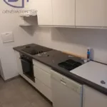 Cuisine équipée dans un style sobre et fonctionnel. Les armoires de cuisine sont blanches avec une finition brillante, ce qui donne à la cuisine un aspect propre et lumineux. En bas, il y a un lave-vaisselle intégré, ce qui est pratique pour la propreté dans la cuisine. À côté, on trouve un four encastré au niveau de la taille, pour une utilisation confortable sans avoir à se pencher. Le plan de travail présente une finition en pierre ou en composite gris, qui contraste avec la blancheur des armoires. Sur ce plan, on distingue une plaque de cuisson à induction moderne avec un espace libre à côté, destiné à préparer les aliments ou à poser des ustensiles de cuisine. Au-dessus du plan de travail, il y a des éléments muraux blancs offrant davantage de rangement. L'un des placards est ouvert, révélant des étagères vides prêtes à être utilisées.