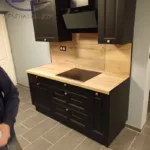 Cette image montre une partie d'une cuisine moderne. Au centre, il y a un plan de travail en bois clair, qui a l'air robuste et fonctionnel, intégrant un évier en inox et une plaque de cuisson. Juste au-dessus, il y a une crédence qui semble être faite dans le même bois que le plan de travail, ajoutant une touche chaleureuse et cohérente à l'ensemble. Les éléments bas de la cuisine sont de couleur noire avec des poignées dorées, ce qui crée un contraste élégant avec le plan de travail en bois clair. Il y a plusieurs tiroirs et placards qui offrent probablement un bon espace de rangement. Sur le côté droit, on voit une armoire haute de la même couleur noire que les éléments bas, avec une porte fermée qui pourrait dissimuler soit du rangement, soit des appareils encastrés comme un four ou un réfrigérateur. Il y a aussi une hotte aspirante moderne de couleur noire qui semble se fondre avec les meubles. Sur le mur, en dessous des éléments hauts, on note la présence de prises électriques, ce qui est pratique pour l'utilisation d'appareils de cuisine. L'éclairage n'est pas directement visible sur la photo, un bon éclairage, important dans une cuisine. Globalement, cette cuisine dégage une impression de modernité et d'élégance, avec un choix de couleurs et de matériaux qui crée un espace accueillant et fonctionnel.