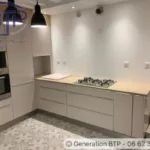 Cuisine moderne et épurée. Les meubles de cuisine sont de couleur blanche avec des poignées discrètes, ce qui donne un aspect moderne à l'ensemble. À gauche, un four encastré et un micro-ondes sont superposés et intégrés dans une colonne de rangement, optimisant l'espace vertical. À côté, il a un espace de rangement ouvert. Le plan de travail est recouvert d'un matériau de protection. Une plaque de cuisson à gaz est installée au centre de ce plan de travail. Trois grandes lampes pendent au-dessus de la cuisine au dessus de la plaque de cuisson, fournissant un éclairage ciblé et un contraste de couleur. À droite, une série de tiroirs sous le plan de travail offre un rangement pratique. Deux spots encastrés de la cuisine.